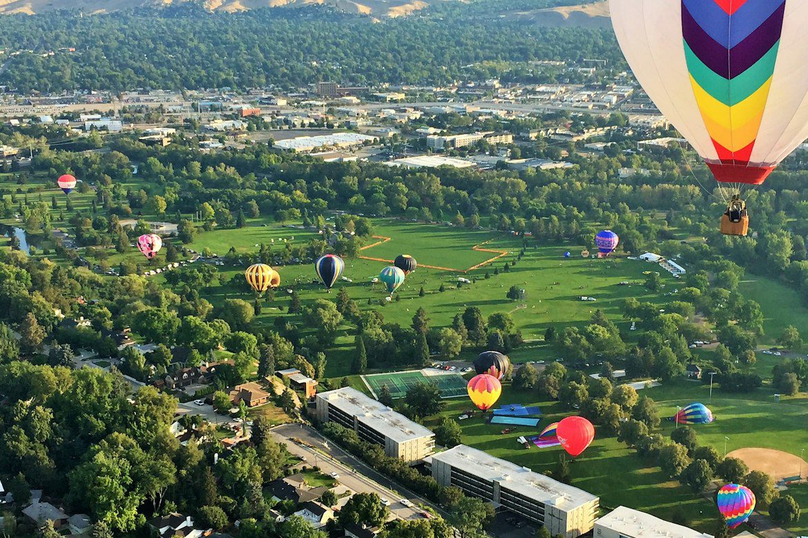 Colorful hot air balloons over a Boise park with tree-lined neighborhoods and foothills beyond